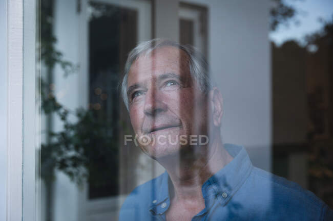 Счастливый пожилой кавказский мужчина в отставке дома смотрит в окно улыбаясь, с отражениями сада в окне, самоизоляция во время коронавируса covid19 пандемии — стоковое фото