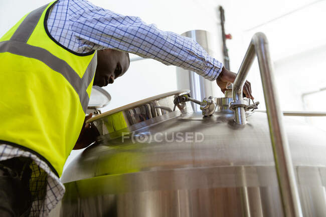 Vue en angle bas d'un homme afro-américain travaillant dans une microbrasserie, portant un gilet haute visibilité, inspectant la bière et regardant à l'intérieur du réservoir. — Photo de stock