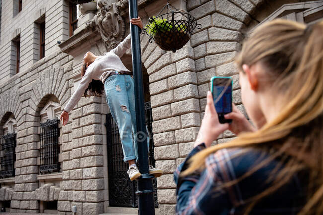 Vue arrière d'un Caucasien et d'une fille métissée profitant du temps passé ensemble par une journée ensoleillée, fille debout sur le trottoir et prenant une photo de son ami debout sur le lampadaire. — Photo de stock
