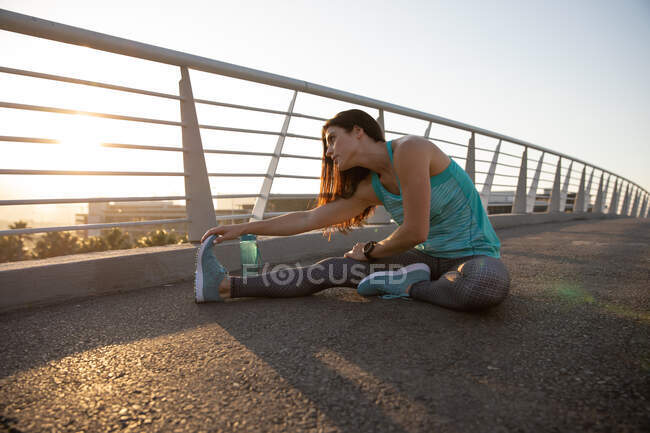 Side view of a fit Caucasian woman with long dark hair wearing sportswear exercising outdoors in the city on a sunny day with blue sky, warming up, sitting and stretching her leg, — Stock Photo