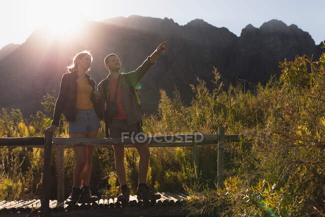 Вид спереди на кавказскую пару, хорошо проводящую время в поездке в горы, стоящую на деревянном мосту, мужчина указывает на что-то, в солнечный день — стоковое фото