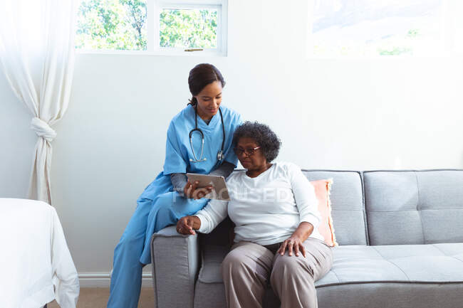 Femme âgée métisse passant du temps à la maison, visitée par une infirmière métisse, utilisant une tablette — Photo de stock