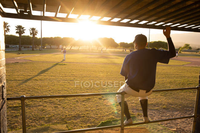 Vista posteriore di un giocatore di baseball maschile di razza mista, guardando una partita di baseball, seduto su una barriera, in una giornata di sole — Foto stock