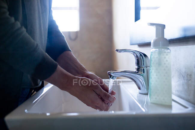 Средняя часть женщины дома в ванной в дневное время моет руки в раковине, бутылка с жидким мылом рядом с ней, защита от коронавируса Covid-19 инфекции и пандемии. Социальное дистанцирование и самоизоляция в карантинной изоляции — стоковое фото