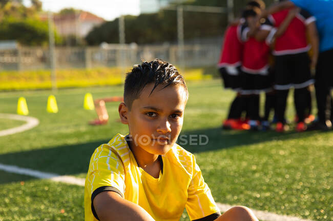 Портрет змішаної раси хлопчика-футболіста, який сидить на ігровому полі на сонці під час тренувальної сесії, дивлячись на камеру і посміхаючись, з його товаришами по команді слухаючи їх тренер на задньому плані — стокове фото