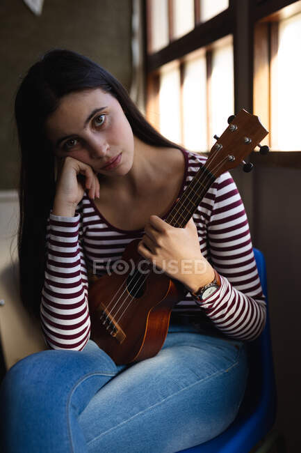 Portrait of a Caucasian musician teenage girl sitting by a window, looking to camera and holding a ukulele in a high school — Stock Photo