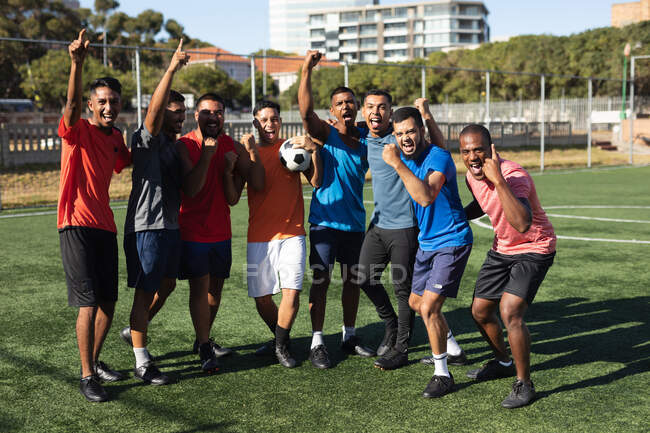 Multi grupo étnico de hombres cinco jugadores de fútbol de lado que usan ropa deportiva de entrenamiento en un campo de deportes en el sol, celebrando la victoria sosteniendo una pelota. - foto de stock