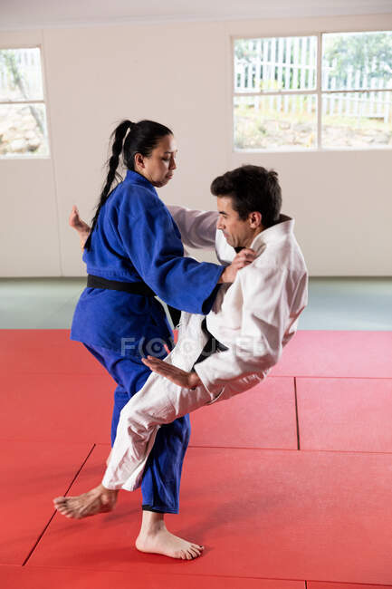 Vista laterale di un allenatore di judo maschile di razza mista e judoka femminile di razza mista adolescente, con indosso judogi blu e bianchi, che pratica il judo durante un allenamento in palestra. — Foto stock