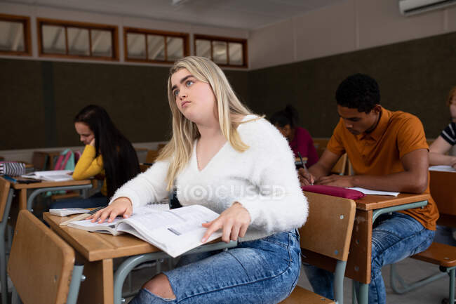 Side view of a teenage Caucasian girl in a school classroom sitting at desk, concentrating, with teenage male and female classmates sitting at desks working in the background — Stock Photo