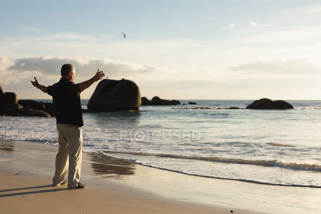 Vista laterale di un anziano caucasico in piedi sulla spiaggia in riva al mare ammirando una vista costiera con le braccia tese in celebrazione — Foto stock