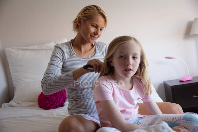 Vue de face d'une femme caucasienne profitant du temps en famille avec sa fille à la maison ensemble, la mère brossant les cheveux de sa fille assise sur un lit dans leur chambre — Photo de stock