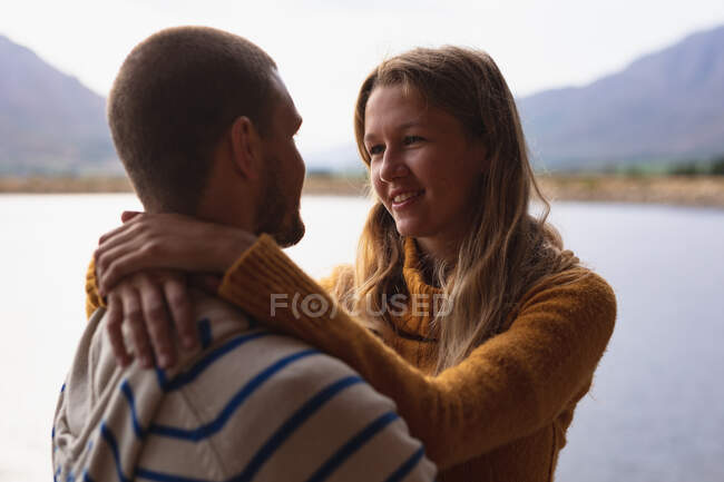 Vista lateral de um casal caucasiano se divertindo em uma viagem às montanhas, de pé em uma varanda em uma cabine, abraçando, olhando um para o outro — Fotografia de Stock