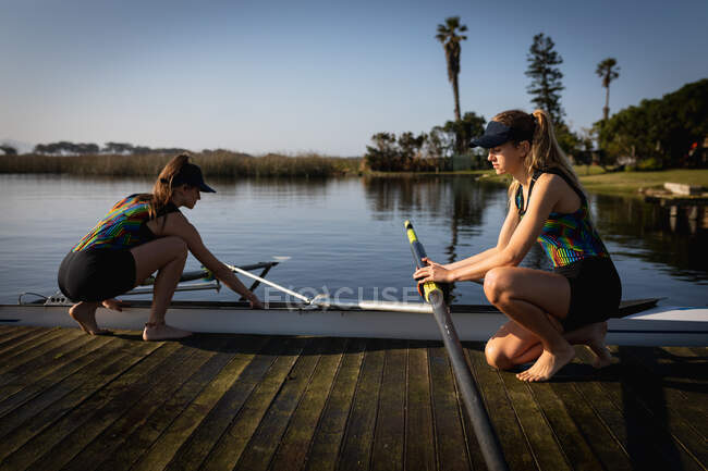 Vista lateral de dos mujeres remadoras caucásicas desde un equipo de remo entrenando en el río, arrodillándose en un embarcadero y preparando un bote en el agua bajo el sol - foto de stock