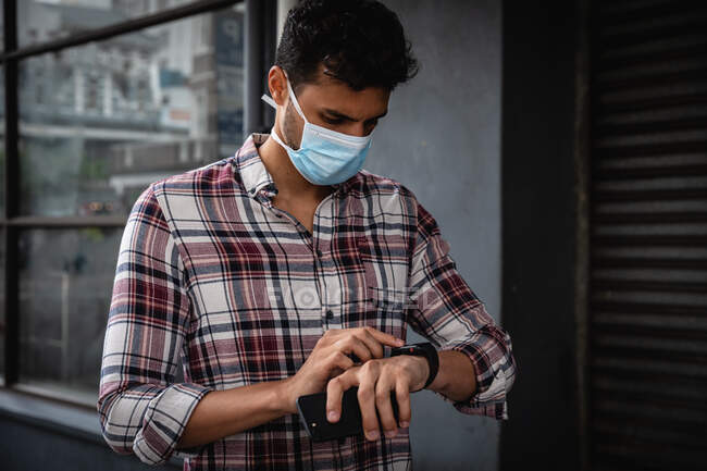 Front view close up of a caucasian man wearing checkered shirt and face mask against air pollution and covid19 coronavirus, walking through the city streets, using his smartwatch. — Stock Photo