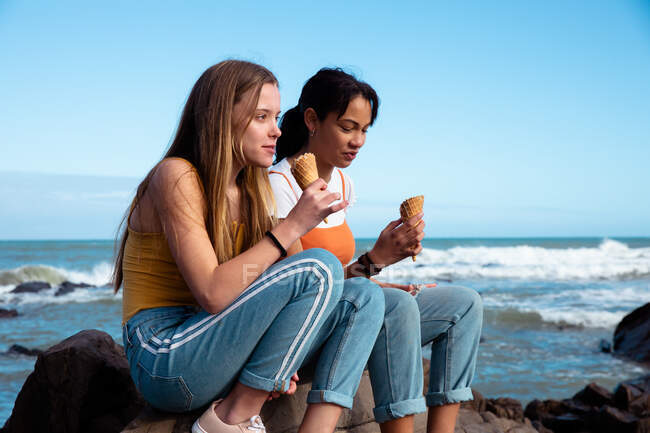 Seitenansicht einer kaukasischen und einer gemischten Rasse Mädchen genießen die Zeit zusammen hängen an einem sonnigen Tag, Eis essen, auf einem Felsen am Strand sitzen. — Stockfoto