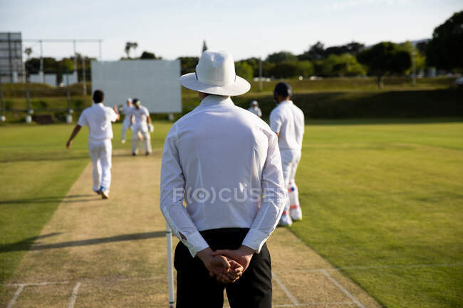Vista posteriore di un arbitro di cricket maschio caucasico che indossa una camicia bianca e un cappello a tesa larga, in piedi su un campo di cricket vicino al wicket, guardando i giocatori durante la partita. — Foto stock