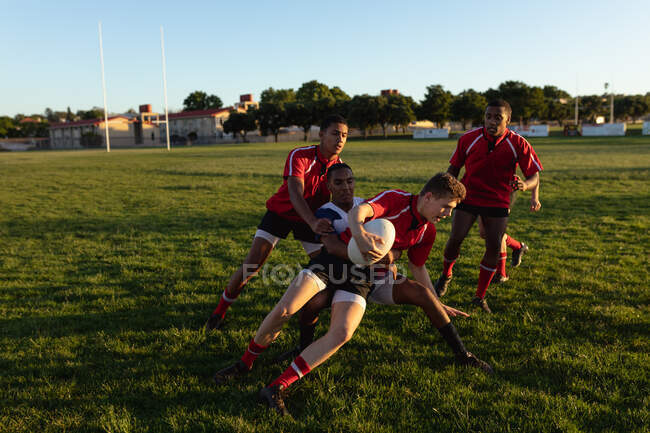 Vista frontal de duas equipes masculinas multiétnicas adolescentes de jogadores de rugby usando suas tiras de equipe, em ação durante uma partida de rugby em um campo de jogo, um jogador em primeiro plano na posse da bola — Fotografia de Stock
