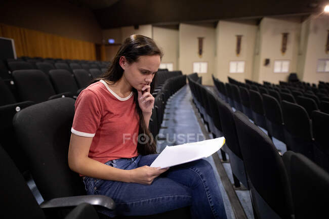 Seitenansicht eines kaukasischen Teenagers in einem leeren Theater der High School, der in der Aula sitzt und sich auf eine Aufführung vorbereitet, ein Drehbuch in der Hand hält und Zeilen lernt — Stockfoto