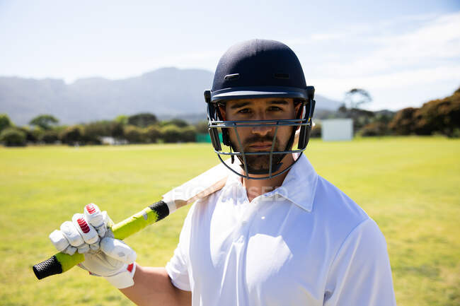 Retrato de um jogador de críquete misto confiante vestindo brancos de críquete, capacete e morcego de críquete, de pé em um campo de críquete em um dia ensolarado olhando para a câmera — Fotografia de Stock