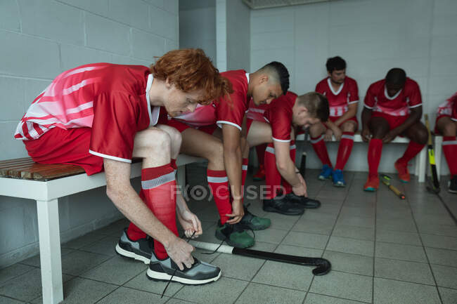 Vista laterale di un gruppo multietnico di giocatori di hockey su prato maschi adolescenti, che si preparano prima di una partita, si siedono nello spogliatoio, legano le scarpe e si concentrano — Foto stock