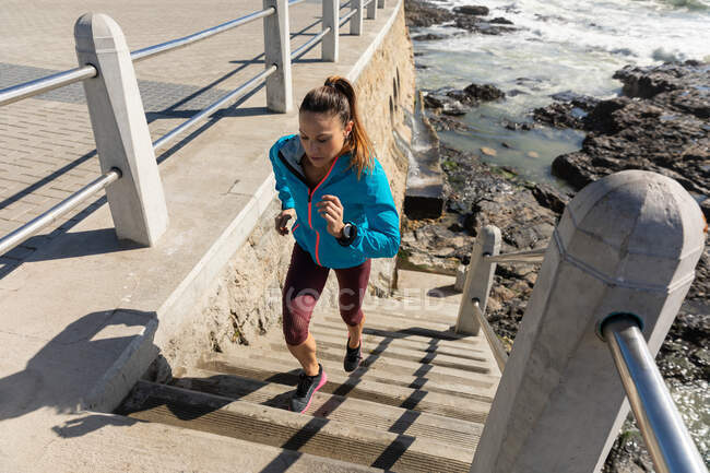 Vista laterale ad alto angolo di una donna caucasica sportiva con lunghi capelli scuri che si esercita su una passeggiata al mare in una giornata di sole, correndo per le scale. — Foto stock