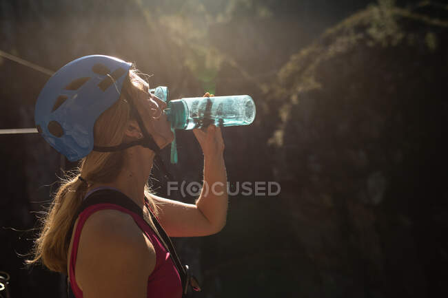 Vista lateral de la mujer caucásica disfrutando del tiempo en la naturaleza, usando equipo de forro de cremallera, poniéndose el casco, agua potable, en un día soleado en las montañas - foto de stock