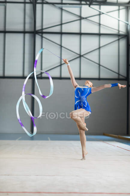 Vista frontal da adolescente caucasiana ginasta feminina realizando no ginásio, exercitando com fita, de pé em uma perna, um braço estendido, olhando para a câmera, vestindo collant azul — Fotografia de Stock