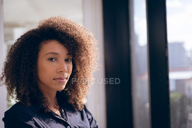 Porträt einer Geschäftsfrau mit gemischter Rasse, die an einem sonnigen Tag in einem modernen Büro neben einem Fenster steht und in die Kamera blickt — Stockfoto