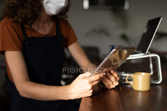Mujer caucásica pasando tiempo en casa, trabajando desde casa, usando su tableta, usando una máscara facial. Estilo de vida en el hogar aislado en cuarentena durante la pandemia del coronavirus covid 19. - foto de stock