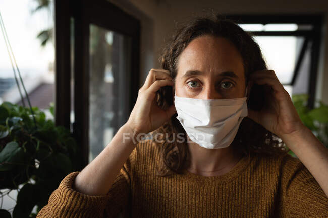Porträt einer kaukasischen Frau, die während der Coronavirus-Covid-19-Epidemie Zeit zu Hause mit Selbstisolierung und sozialer Distanzierung in Quarantäne verbringt, eine Gesichtsmaske gegen Covid19-Coronavirus aufsetzt und direkt in eine Kamera blickt. — Stockfoto