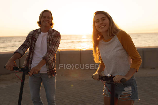 Portrait of a Caucasian couple riding scooters on a promenade during sunset, looking at the camera, relaxing during an active seaside beach holiday — Stock Photo