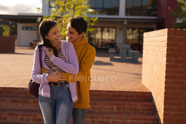 Front view of a Caucasian teenage girl and boy embracing and smiling at each other standing in their school grounds — Stock Photo