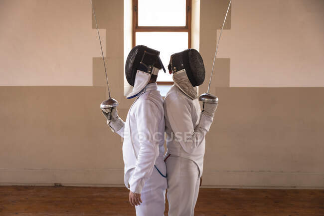 Caucasian and a mixed race sportsmen wearing protective fencing outfit during a fencing training session, standing back to back, holding epees in front of faces. Fencers training at a gym. — Stock Photo