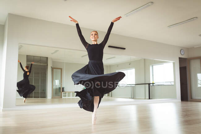 Bailarina de ballet femenina atractiva caucásica con baile de pelo rojo, vestida con un vestido largo y negro, preparándose para una clase de ballet en un estudio brillante, enfocándose en su ejercicio, sonriendo. - foto de stock