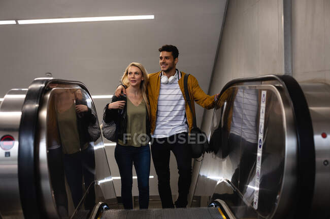 Vista frontal de una pareja caucásica en la ciudad, subiendo en la estación de metro con una escalera mecánica, sonriendo y abrazando. - foto de stock
