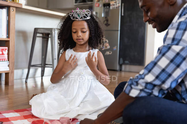 Ragazza afroamericana che indossa una corona giocattolo, distanza sociale a casa durante il blocco di quarantena, giocare con suo padre, avere un tea party bambola. — Foto stock