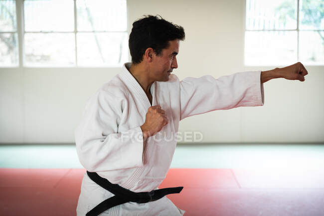 Vista lateral de un entrenador de judo masculino de raza mixta que usa judogi blanco, calentándose antes de un entrenamiento en un gimnasio, posando, golpeando el aire. - foto de stock