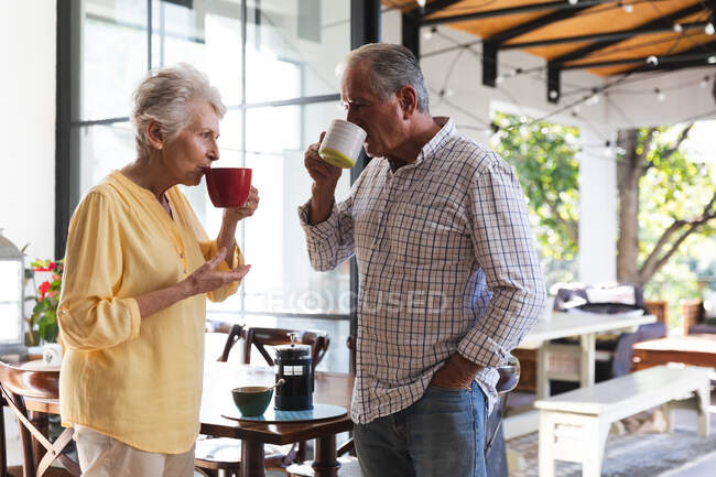 Посторонний вид пожилой белой пары в доме, стоящей на кухне, разговаривающей и пьющей кофе вместе в солнечный день, пары, изолирующей во время пандемии коронавируса — стоковое фото