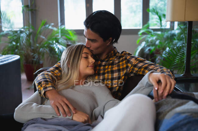 Vista frontale da vicino di un giovane uomo di razza mista e una giovane donna caucasica che si godono il tempo a casa, si siedono nel loro soggiorno e abbracciano, l'uomo bacia la donna sulla fronte. — Foto stock