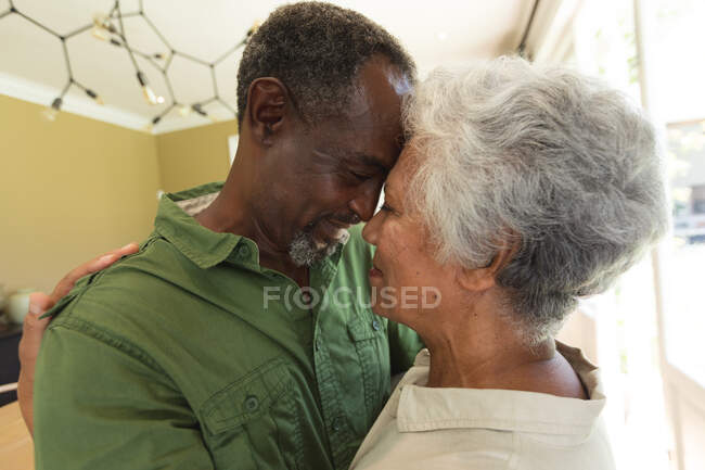 Зіткнення щасливої пари афроамериканців у відставці, які стоять вдома на кухні, торкаючись голів разом, дивлячись один на одного і посміхаючись, обіймаючи, вдома, ізолюючись під час коронавірусної ковірусної ковідемії. — стокове фото