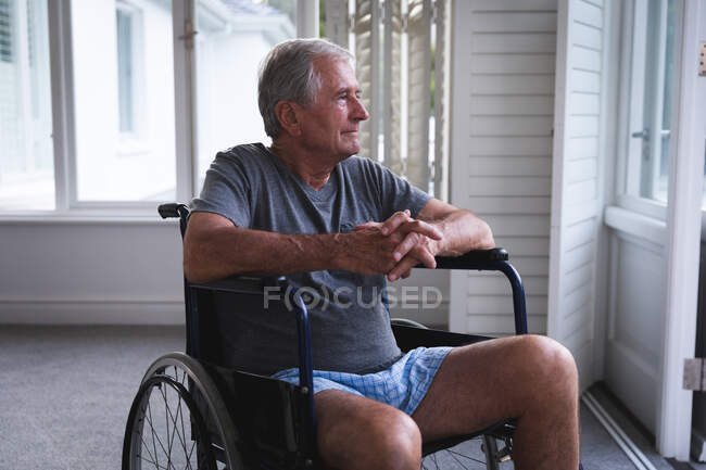 Вийшов на пенсію старший кавказький чоловік вдома, сидячи в інвалідному візку в білизні перед вікном, в сонячний день, озираючись від думки, самоізолюючись під час коронавірусної ковідемії — стокове фото