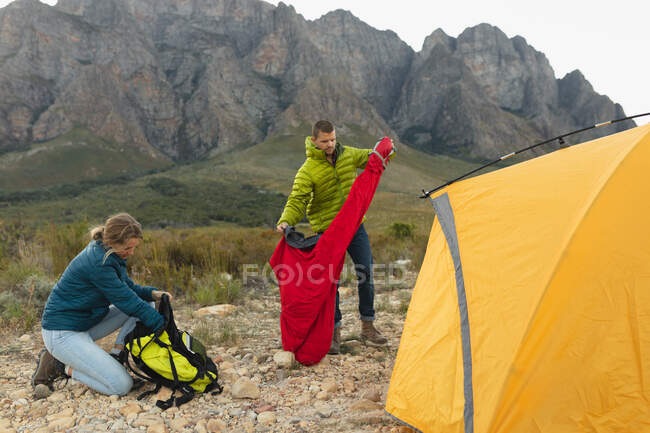 Vista lateral de um casal caucasiano se divertindo em uma viagem às montanhas, em pé perto de uma tenda, tirando coisas de suas mochilas — Fotografia de Stock