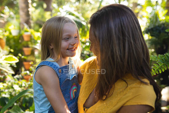 Кавказька жінка з дочкою проводять час разом у сонячному саду, обіймаючи, дивлячись один на одного і посміхаючись — стокове фото