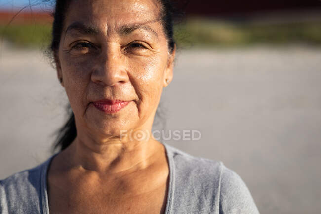 Портрет змішаної старшої жінки, що носить сіру сорочку, стоїть на сонячному пляжі, дивиться прямо на камеру і посміхається . — стокове фото
