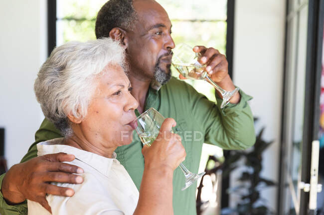 Щаслива високопоставлена подружня пара афроамериканців у відставці вдома, обнімаючи і випиваючи келихи білого вина, подружжя вдома ізольоване під час коронавірусної ковідемії. — стокове фото