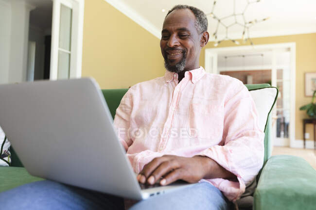Feliz anciano apuesto jubilado afroamericano en casa sentado en un sillón en su sala de estar, utilizando un ordenador portátil y sonriendo, auto aislante durante coronavirus covid19 pandemia - foto de stock