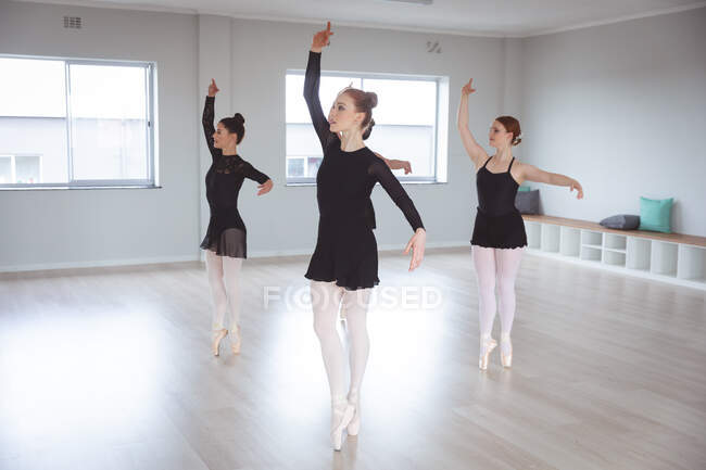 Un groupe de jolies danseuses de ballet caucasiennes en tenues noires, collants blancs et chaussures pointes pratiquant lors d'un cours de ballet dans un studio lumineux — Photo de stock