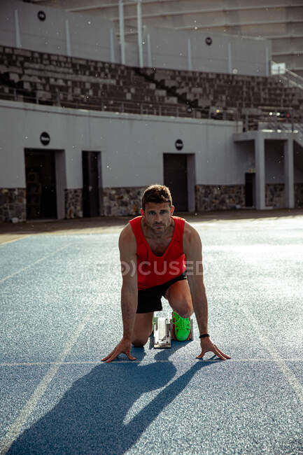 Vista frontale di un atleta caucasico di sesso maschile che pratica in uno stadio sportivo, in posizione sui blocchi di partenza, preparandosi a correre, con la testa alzata — Foto stock