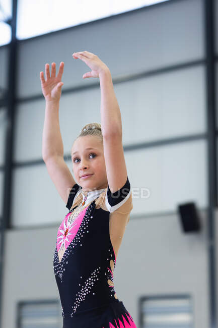SIde view close up of happy teenage Caucasian female gymnast performing at sports hall, standing with arms up, wearing pink, black and beige leotard — Stock Photo