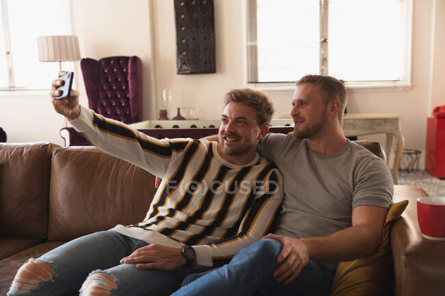 Front view of Caucasian male couple relaxing at home, sitting on a sofa, embracing, smiling and taking selfie with their smartphone — Stock Photo